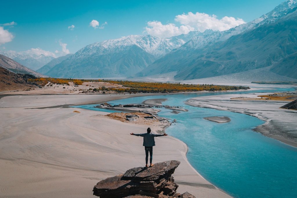 This is the image of a person who travels, standing on top of a rock, in a scenery or a river with mountains in the background, extending their arms to the uncertainty and beauty of traveling. 